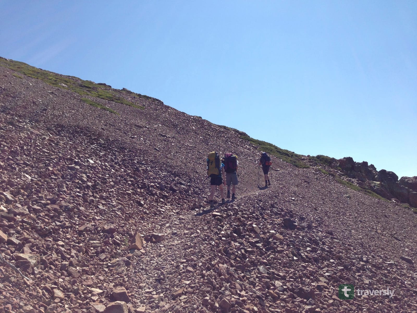 Three hikers with loaded backpacks climbing a loose scree slope on the GR20.