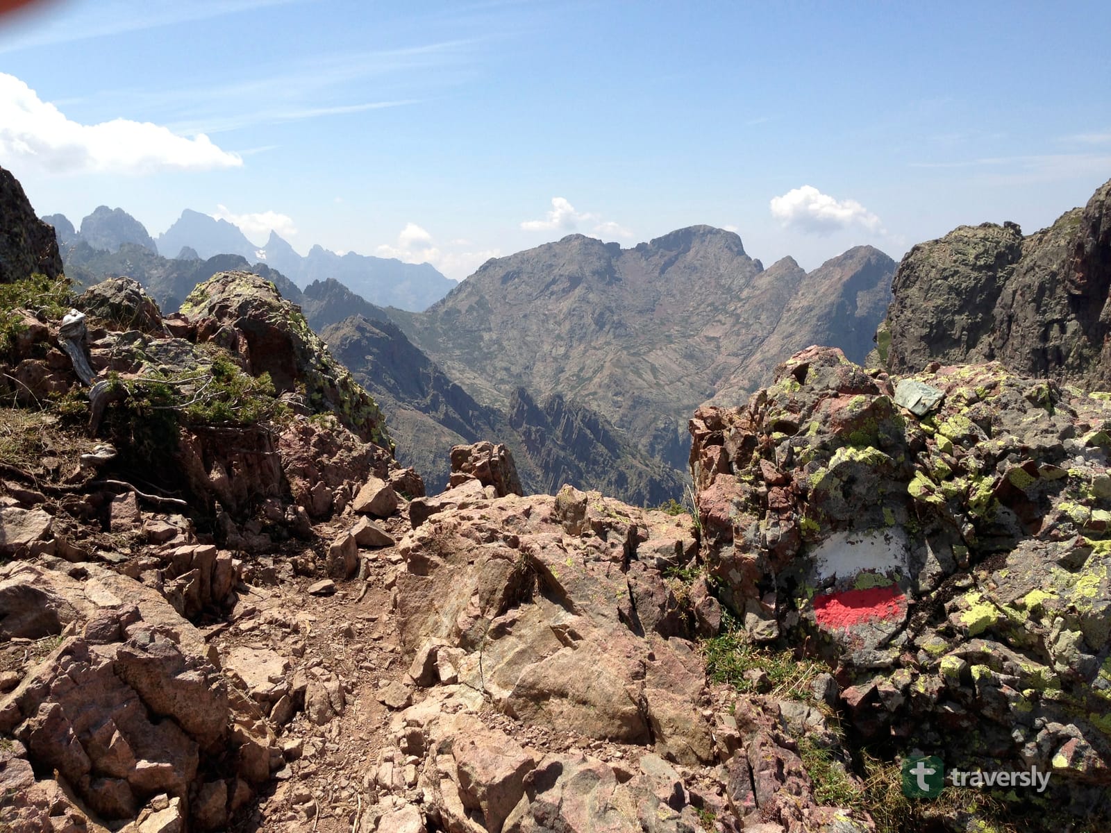 A red-and-white GR20 waymark painted on a granite outcrop, with Corsica's jagged interior peaks in the background.